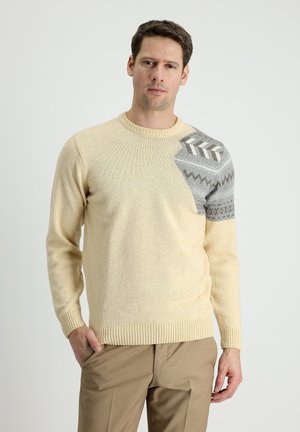 Beige Strickpullover mit Rundhalsausschnitt; verfügt über ein graues gemustertes Patch auf einer Schulter mit geometrischen Designs. Rippbündchen und Saum.