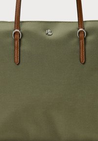 Olivgrön canvas tote-väska med bruna läderhandtag och silverdetaljer. Har präglad logotyp i mitten, slät textur.