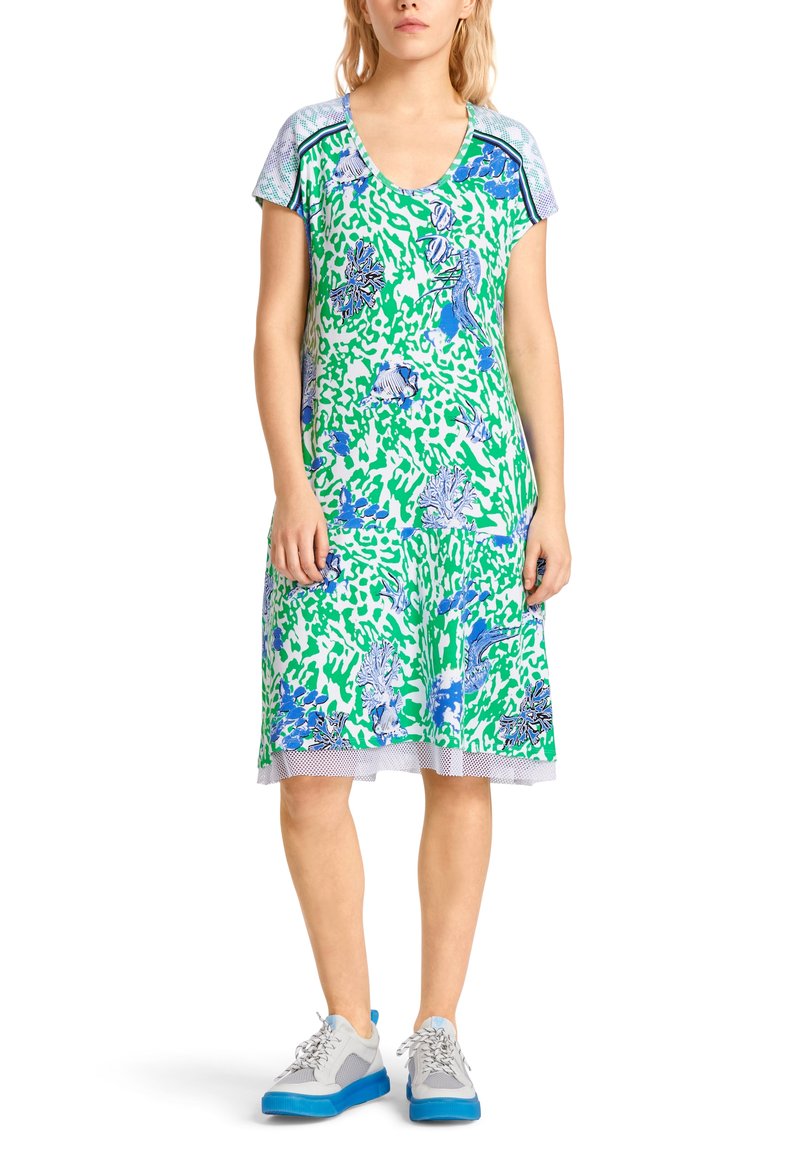 Femme portant une robe verte et blanche longueur genoux avec des motifs floraux et poissons bleus, associée à des baskets blanches et bleues.