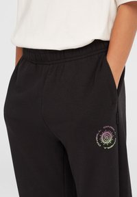 Svarta joggingbyxor med elastisk midja, sidofickor och en flerfärgad cirkulär logotyp. Slät textur och avslappnad passform.