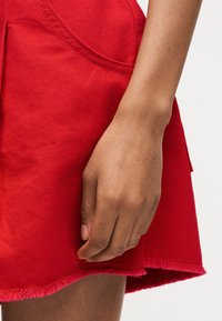 Δεξί χέρι που ακουμπάει δίπλα σε κόκκινη φούστα από τζιν με φθαρμένο τελείωμα και εμφανές ράψιμο στις τσέπες.