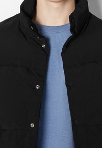 Černá vyplněná bunda s vysokým límcem, druky a texturovaným povrchem, vrstvená přes světle modrý svetr.