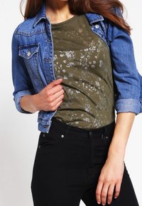 Jeansjacka med en ljusblå tvätt, lagd över en olivgrön T-shirt med blommönster. Svarta jeans fulländar outfiten.