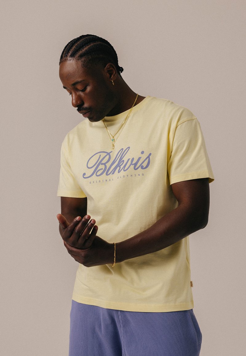 Homme aux cheveux tressés portant un t-shirt jaune « Blkvis Original Clothing » et un pantalon bleu, réglant son poignet gauche avec sa main droite.
