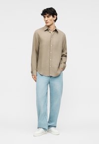 Νεαρός άνδρας ντυμένος με μπεζ πουκάμισο με κουμπιά, ανοιχτόχρωμο τζιν με φαρδιά πόδια και λευκά αθλητικά παπούτσια, στέκεται με το ένα χέρι στην τσέπη.