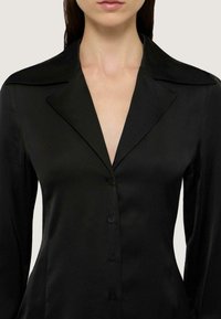 Camicia in satin nero con scollo a V profondo, colletto affilato e maniche lunghe con chiusura a bottoni. Texture liscia e vestibilità su misura.