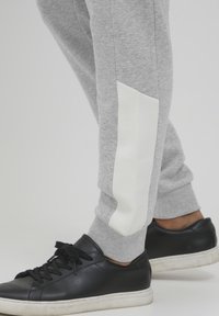 Graue Jogginghose aus weichem Stoff mit einem weißen Akzent an der Seite. Kombiniert mit schwarzen Low-Top-Sneakern aus strukturiertem Leder.