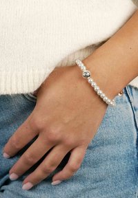 Elastiskt armband med vita pärlor och en silverpärla. Slät yta, rund form, bärs på en hand mot en ljus tröja och jeans.