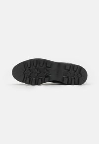 Black rubber shoe sole with deep treads, featuring a textured surface and angular patterns for enhanced grip and stability.