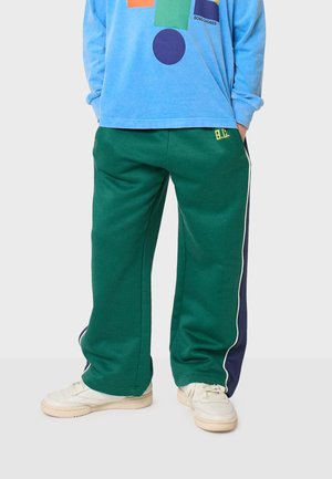 Grüne Sweatpants mit weißem Seitenstreifen, elastischem Bund und kleinen gelben Stickdetails. Tragen kombiniert mit einem blauen Langarmshirt und Turnschuhen.