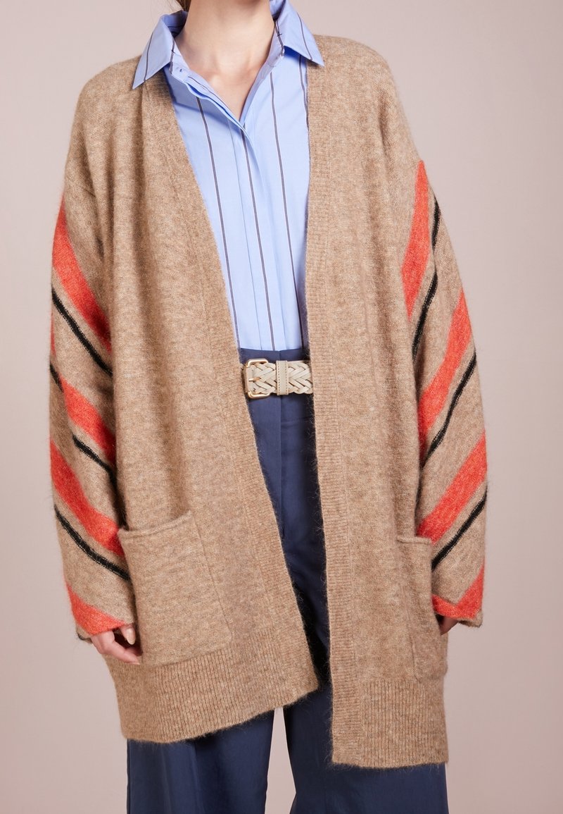 Femme portant un cardigan beige surdimensionné avec des rayures rouges et noires sur les manches, une chemise à rayures bleues, un pantalon bleu marine et une ceinture tressée.