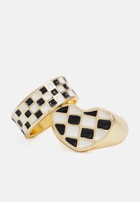 Dois anéis: um apresenta uma forma de coração redonda com um padrão quadriculado em preto e branco, enquanto o outro possui uma faixa com um design de grelha e detalhes em dourado.