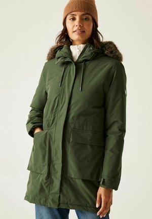 Kobieta w zielonej zimowej kurtce parka z futrzanym kapturem, białym swetrze na zamek, niebieskich dżinsach i brązowej dzianinowej czapce.