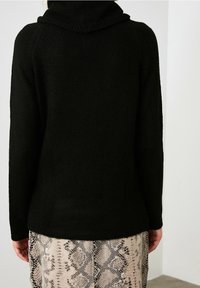 Eine Person trägt einen schwarzen Pullover mit langen Ärmeln und einen beigen Rock mit Schlangenprint und steht vor einer einfarbigen, hellen Wand.