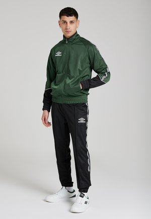 Giacca a track in nylon verde con maniche nere, chiusura a zip e logo. Pantaloni jogger neri con logo bianco e strisce laterali a motivo. Sneakers bianche.