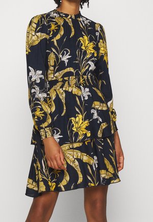 Femme portant une robe bleu marine à manches longues avec des motifs floraux et de feuilles jaunes et blanches, taille élastique et couches de jupe à volants.