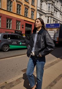 Svart läderbombarjacka med blå krage, kombinerad med en ljusrandig tröja och blå jeans, bärs på en stadsgata.