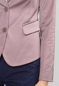 Blazer rose poussiéreux avec boutons et poche à rabat porté sur un pantalon bleu marine foncé, montrant la partie médiane du corps et le bras droit.
