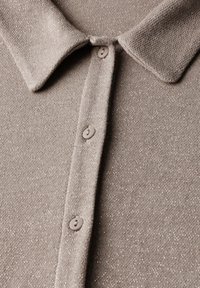 Polo en tricot beige avec un subtil pailleté, présentant un col classique et trois boutons sur le devant. Texture douce et tissu côtelé.