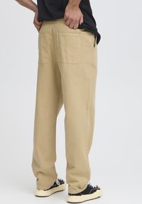 Pantalon en coton beige à coupe décontractée, doté d'une ceinture élastique, de deux poches arrière, porté avec des baskets noires.