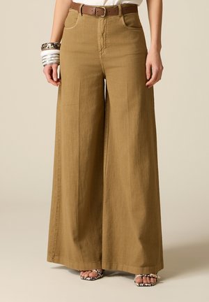 Hnědé kalhoty s vysokým pasem a širokými nohavicemi v kombinaci s hnědým opaskem, bílým topem, sandály se zvířecím potiskem a náramky na levém zápěstí.