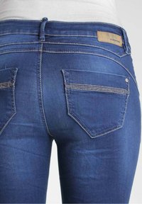 Smal blå jeans i denim, med fem fickor, kontrasterande sömmar och en etikett på midjebandet. Texturen är slät med en lätt glans.