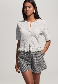 Witte kanten cardigan met korte mouwen, voorzien van een knoopsluiting aan de voorkant en een gerimpelde zoom. Gecombineerd met zwart-wit geruitte shorts.