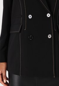 Blazer nero con cuciture beige a contrasto, design doppiopetto, due tasche frontali e grandi bottoni rotondi lucidi neri e argento.