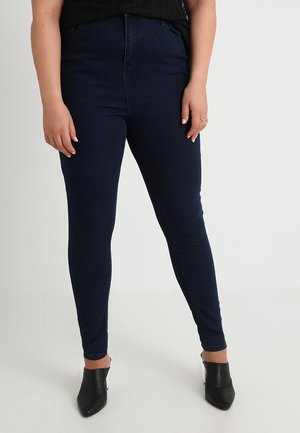 Henkilö, joka pukeutunut tummansinisiin kapeisiin farkkuihin, mustaan hihattomaan toppiin ja mustiin korkkareihin, joissa on terävä kärki, seisoo yksivärisen vaalean taustan edessä.