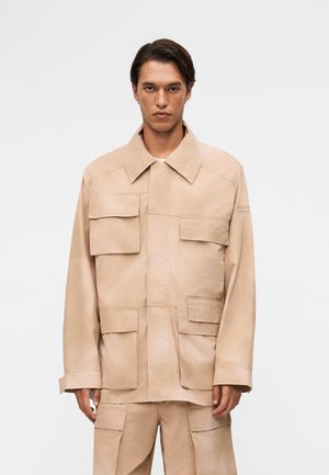 Άνδρας που φοράει μπεζ φαρδιά μπουφάν και ταιριαστό παντελόνι με πολλές μεγάλες τσέπες με καπάκι, στέκεται μπροστά σε απλό ανοιχτό φόντο.