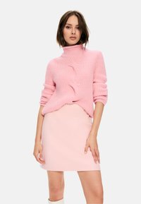 Rosa gestrickter Pullover mit hohem Kragen und geripptem Muster, kombiniert mit einem figurbetonten hellrosa Rock. Das Model posiert mit einer Hand auf der Hüfte.
