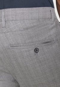 Šedé kalhoty na míru s jemným károvaným vzorem, doplněné černým knoflíkem a bočními kapsami. Vyrobeno z texturovaného materiálu.