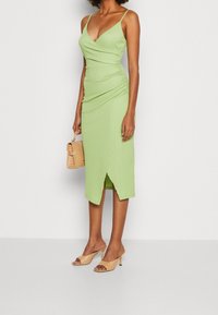 Kvinna i en ärmlös ljusgrön midiklänning med en omslagsskjol, som håller en beige handväska och bär beige sandaler med öppen tå.