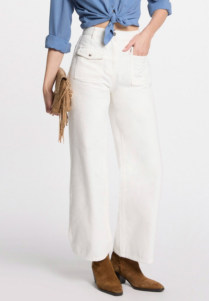 Weiße weit geschnittene Jeans mit Fronttaschen und hoher Taille, kombiniert mit einer blauen gebundenen Bluse und braunen Wildleder-Stiefeletten.