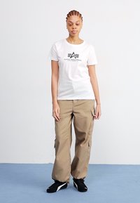 T-shirt blanche avec le logo noir "Alpha Industries", associée à un pantalon cargo beige ample et des sneakers noires sur un sol bleu clair.