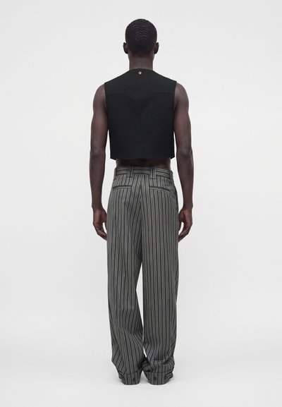 Homme portant un haut court noir sans manches et un pantalon ample gris à fines rayures, debout dos à la caméra sur un fond uni.