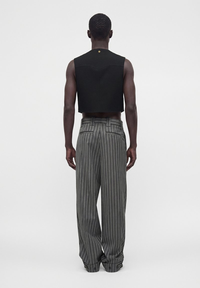 Homme portant un haut court noir sans manches et un pantalon ample gris à fines rayures, debout dos à la caméra sur un fond uni.
