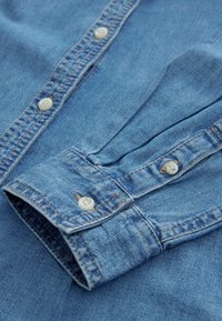 Hellblaues Jeanshemd mit Button-Down-Kragen, weißen Knöpfen und kontrastierenden Nähten. Gefalteter Ärmel zeigt die Manschettendetails.