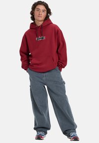 Maroon hoodie with drawstrings and front pocket, paired with loose-fitting grey jeans. Sneakers feature multicolored accents.
