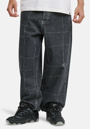 Άνδρας που φοράει φαρδιά μαύρα τζιν με λευκές γραμμές σε σχέδιο πλέγματος και μαύρα αθλητικά παπούτσια DC, στέκεται σε λευκό φόντο.