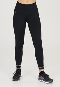 Leggings noirs taille haute avec une texture lisse, des poches en mesh sur les côtés et des accents réfléchissants au niveau de la cheville ; associés à des chaussures de sport noires.
