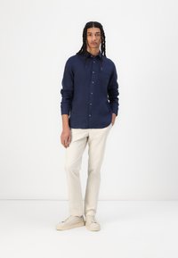 Camicia a maniche lunghe blu navy con chiusura a bottoni, abbinata a pantaloni color crema e scarpe da ginnastica chiare, in piedi su uno sfondo bianco uniforme.