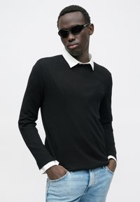 Suéter negro de punto con camisa blanca de cuello debajo. Modelo lleva gafas de sol negras. Vaqueros azul claro y tono de piel oscuro.