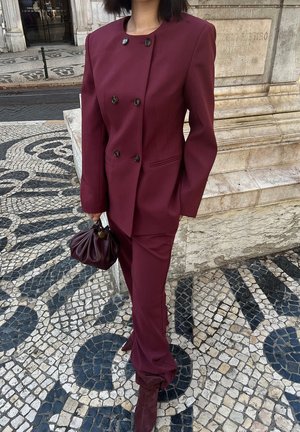 Bordeaux dubbelzijdig pak met opvallende knopen, nauwsluitende pasvorm en bijpassende high-waisted broek, gecombineerd met een geplooide handtas.