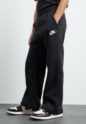 Person iført sorte Nike sweatpants med hænderne i lommerne og sorte og hvide sneakers stående på en grå gulv.