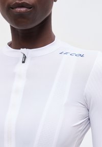 Gros plan sur une personne portant un haut blanc zippé avec le logo "LE COL" sur l'épaule, mettant en valeur l'encolure et la partie supérieure de la poitrine.