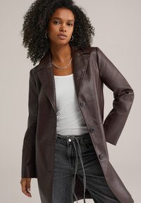 Abrigo largo de cuero marrón con textura suave, solapas con muesca, dos botones y bolsillos frontales, combinado con una camisa blanca y jeans oscuros.