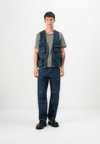 Denimweste mit mehreren Taschen, kombiniert mit einem grauen T-Shirt und dunklen Jeans. Schwarze Schuhe mit dicker Sohle runden das Outfit ab.