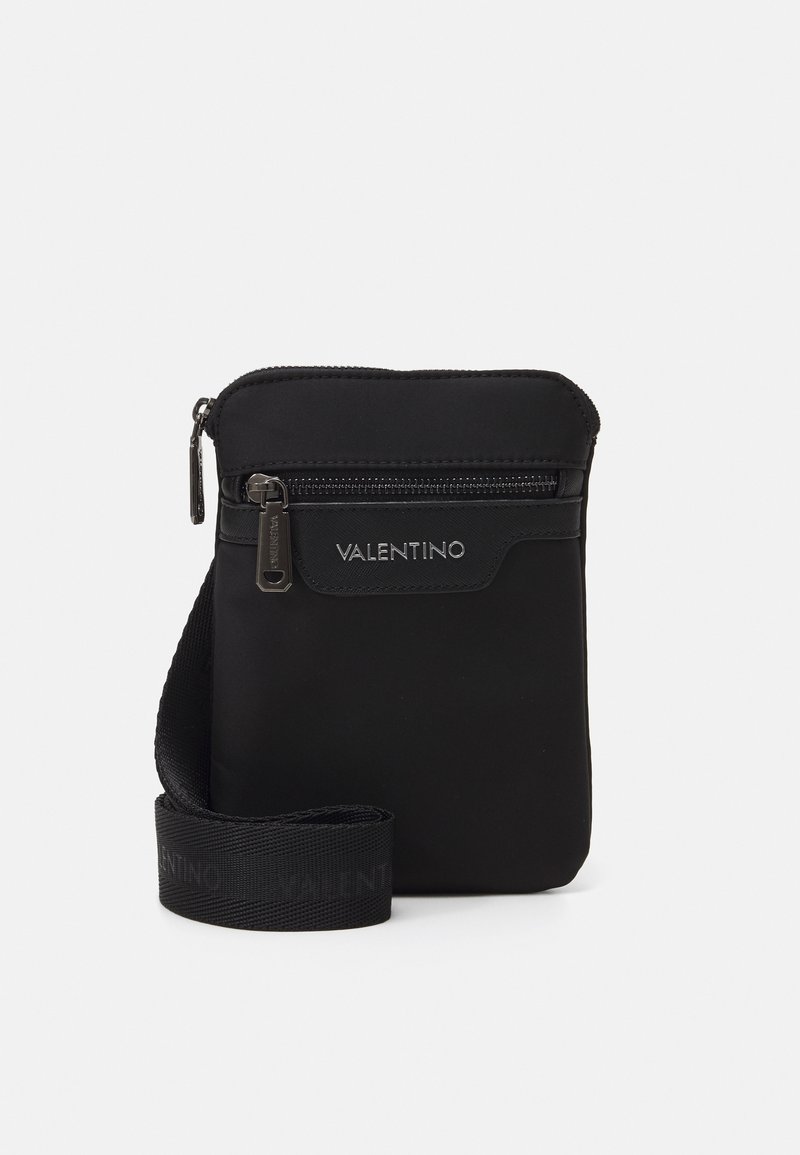 Sac à épaule en nylon noir avec une poche avant zippée, une sangle réglable et un branding discret. Texture lisse et design minimaliste.