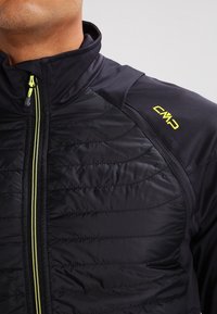 Sort jakke med quiltet front, der har høj krave, gul lynlås og logo på skulderen. Lavet af teksturede, letvægtsmaterialer.